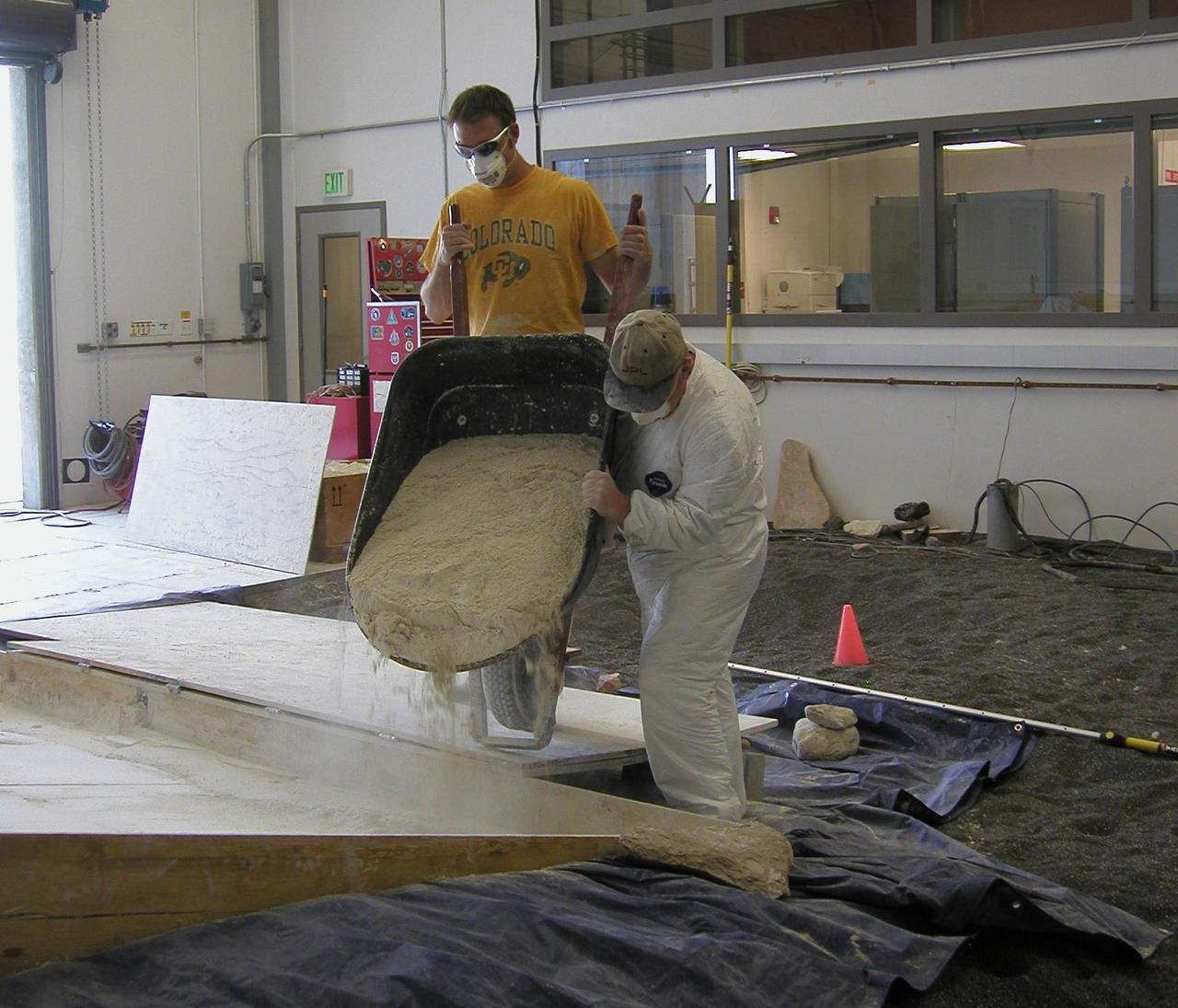 Rover team members Mike Seibert left and Paolo Bellutta add a barrowful of soil mixture to the sloped box where a test rover will be used for assessing possible maneuvers for NASA rover Spirit to use in escaping from a sandtrap on Mars.