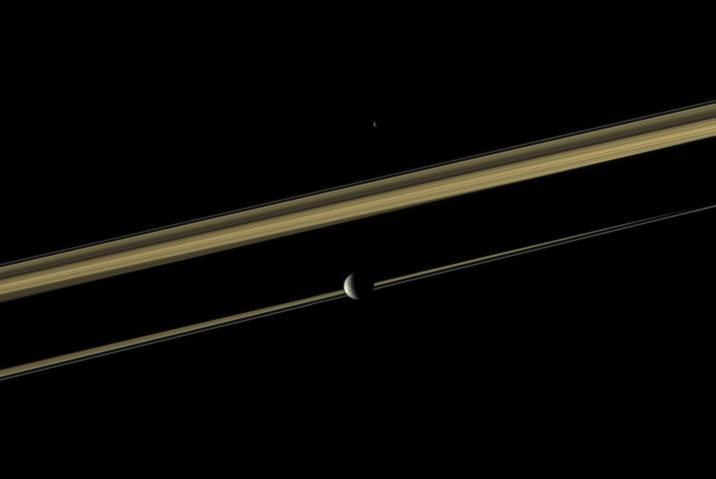 Two of Saturn moons straddle the planet rings in this color view from NASA Cassini spacecraft. Mimas is closest to NASA Cassini spacecraft here. Epimetheus is on the far side of the rings. Saturn shadow cuts across the middle of the rings.