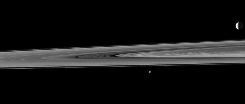 Saturn rings occupy the space between two of the planet moons in this image, taken by NASA Cassini spacecraft, which shows the highly reflective moon Enceladus in the background and the smaller moon Janus in the fore.