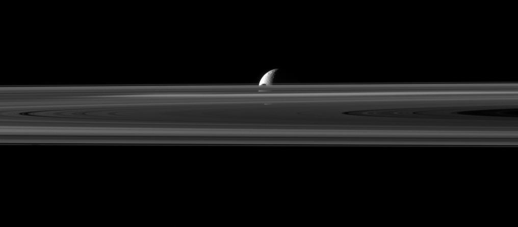 The small moon Janus is almost hidden between the planet rings and the larger moon Rhea in this image captured by NASA Cassini spacecraft. The northern part of Janus can be seen peeking above the rings.
