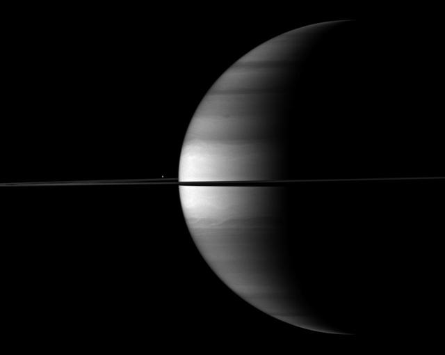 The highly reflective moon Enceladus appears as a bright dot beyond a crescent Saturn in this image from NASA Cassini spacecraft. Enceladus is visible above the ringplane to the left of the center of the image.