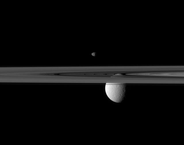 Saturn rings occupy the foreground of this image captured by NASA Cassini spacecraft. The small moon Janus appears to hover above, while the far larger moon Rhea is partially obscured by the rings.