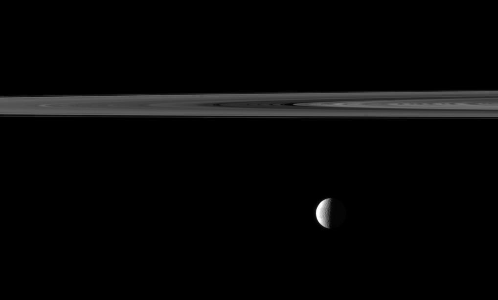 An illuminated quarter of the moon Tethys is imaged near a swath of Saturn rings. Though the moon appears to be hanging directly below the rings, Tethys is actually farther from NASA Cassini spacecraft, and the rings are in the foreground.