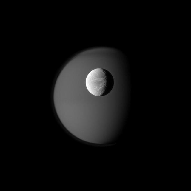 The surface of Saturn moon Dione is rendered in crisp detail against a hazy, ghostly Titan. A portion of the wispy terrain of Dione trailing hemisphere can be seen on the right in this captured by NASA Cassini spacecraft.
