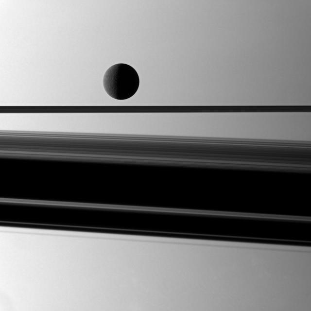 Saturn moon Rhea is gently lit in front of a background of the planet with a wide shadow cast by the rings which are seen nearly edge-on in this image captured by NASA Cassini spacecraft.