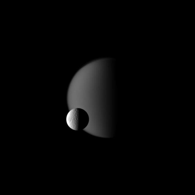 Terrain on Saturn moon Tethys, defined with craters, is shown in front of the hazy atmosphere of the larger moon Titan in this image from NASA Cassini spacecraft. This view looks toward the Saturn-facing sides of Titan and Tethys.