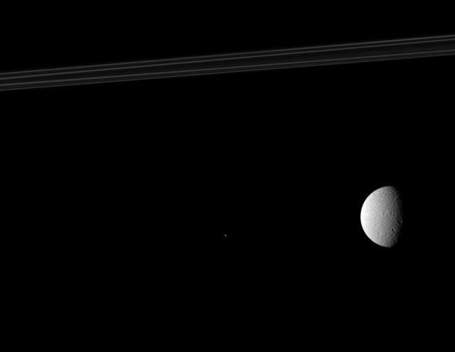 NASA Cassini spacecraft composition features Saturn rings top of image, its second largest moon, Rhea on right of image, and one of the planet tiny moons, Telesto near the middle of image appears as a bright speck.