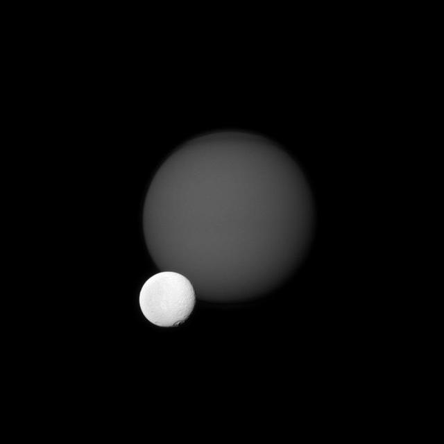 NASA Cassini spacecraft views the cratered surface of Saturn moon Tethys in front of the hazy orb of the planet largest moon, Titan. Tethys is much closer than Titan to Cassini.
