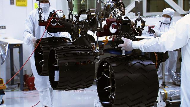 In the middle of this image, three wheels are shown raised by a lift, with engineers on both sides of the wheels in the cleanroom, where NASA Curiosity rover is being assembled.