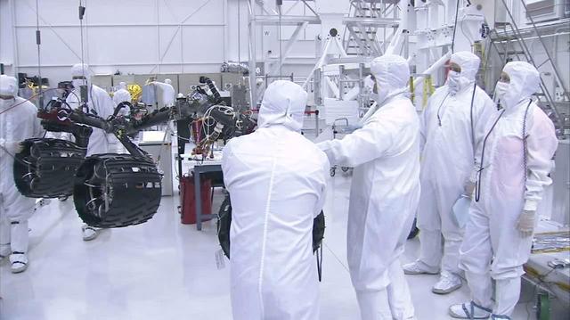 In this image taken from a video clip, NASA Curiosity rover wheels are shown hoisted on a pink crane and moving closer to the rover body. Video is available at the Photojournal.