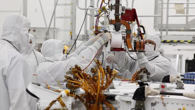 Engineers at NASA Jet Propulsion Laboratory are dressed head to toe in bunny suits only their eyes and foreheads can be seen. They are huddled around the base of the rover Curiosity neck its Mast.