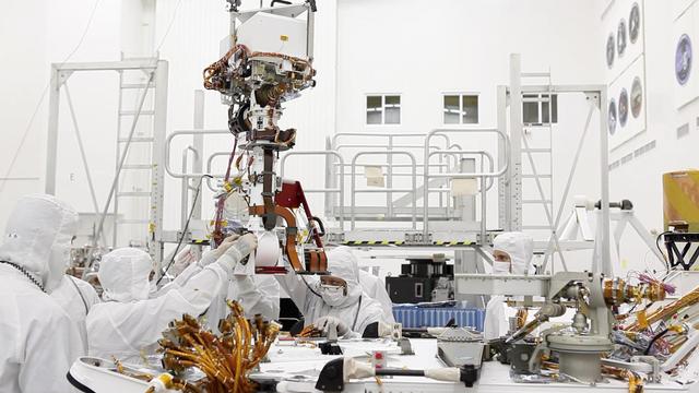 In the clean room at NASA Jet Propulsion Laboratory, engineers gather around the base of Curiosity neck the Mast as they slowly lower it into place for attachment to the rover body the Wet Electronics Box, or WEB.