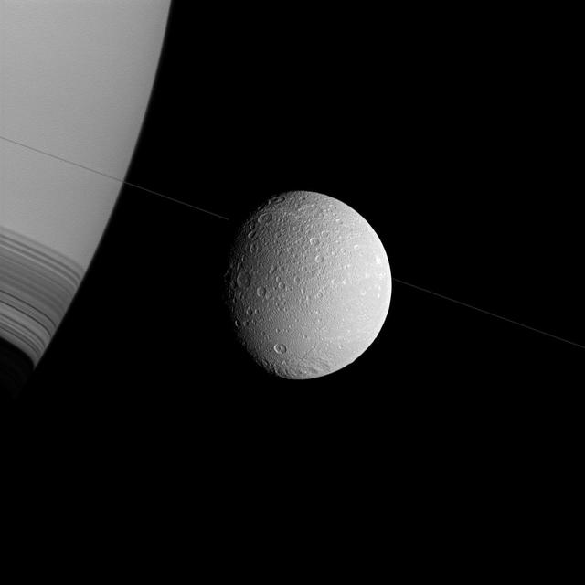 Saturn and Dione appear askew in this view from NASA Cassini spacecraft, with the north poles rotated to the right, as if they were threaded along on the thin diagonal line of the planet rings.