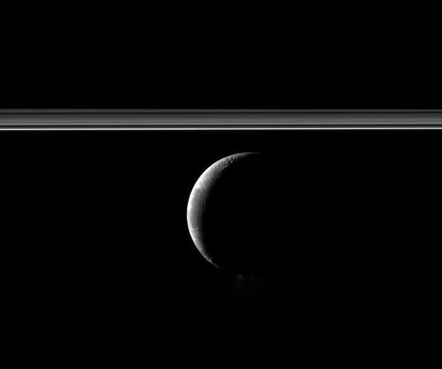 A crescent Enceladus appears with Saturn rings in this view of the moon from NASA Cassini spacecraft. The famed jets of water ice emanating from the south polar region of the moon are faintly visible here.