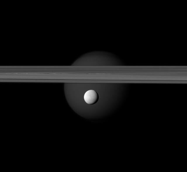 The brightly reflective moon Enceladus appears before Saturn rings while the larger moon Titan looms in the distance. Jets of water ice and vapor emanating from the south pole of Enceladus hinting at subsurface sea rich in organics.