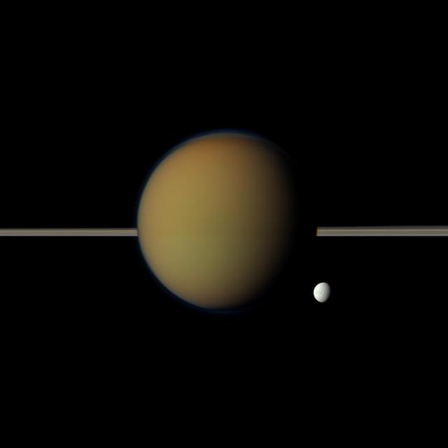 Saturn moon Tethys, with its stark white icy surface, peeps out from behind the larger, hazy, colorful Titan in this view of the two moons obtained by NASA Cassini spacecraft. Saturn rings lie between the two.