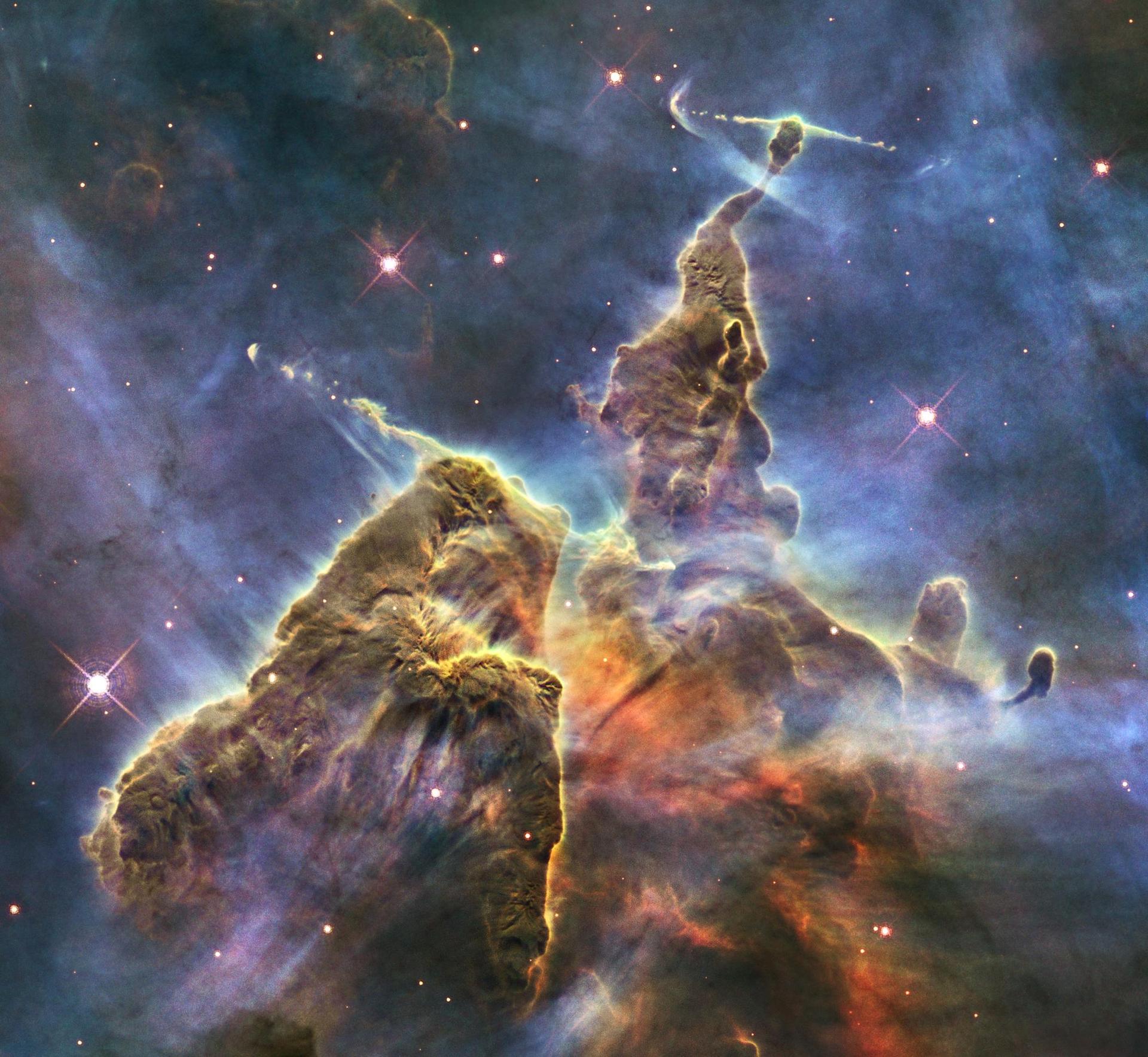 The Mystic Mountain is seen as a chaotic pillar of colorful gas and dust, narrowing toward the top of the image. The dust and gas is mostly yellow, brown, and orange, all jutting against a hazy purple and blue background with a few pink stars.