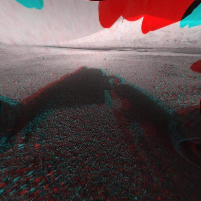This image is a 3-D view in front of NASA Curiosity rover. Mount Sharp is visible rising above the terrain, though in one eye a box on the rover holding the drill bits obscures the view.
