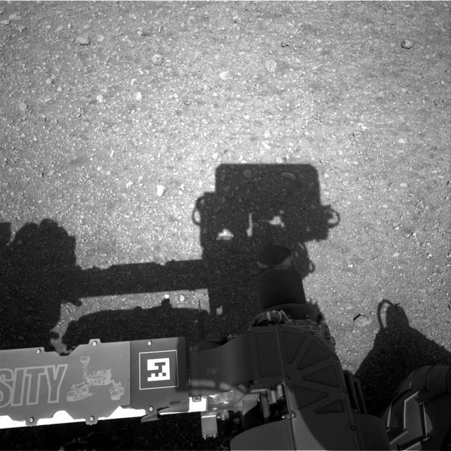 This is the first image taken by the Navigation cameras on NASA Curiosity rover. It shows the shadow of the rover now-upright mast in the center, and the arm shadow at left. The arm itself can be seen in the foreground.