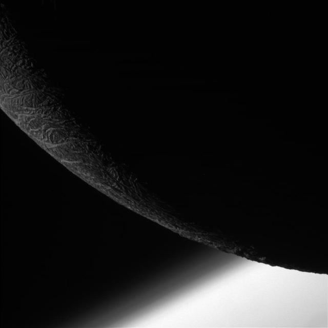 NASA Cassini orbiter shows its final close flyby of Enceladus to focus on the icy moon craggy, dimly lit limb, with the planet Saturn beyond.