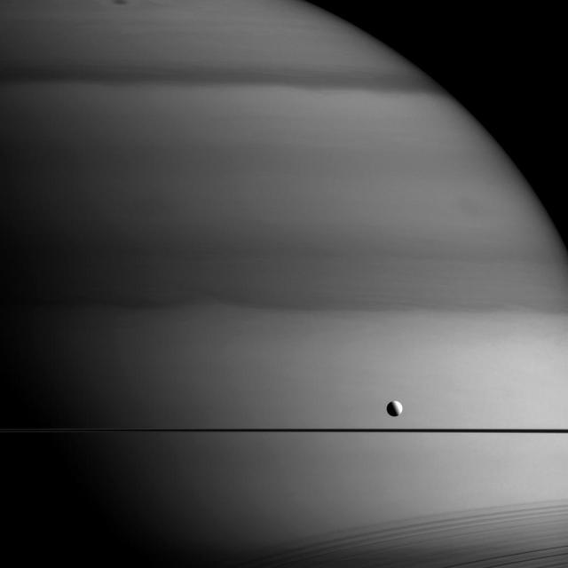 Why does Saturn look like it's been painted with a dark brush in this infrared image, but Dione looks untouched? Perhaps an artist with very specific tastes in palettes?  The answer is methane. This image was taken in a wavelength that is absorbed by methane. Dark areas seen here on Saturn are regions with thicker clouds, where light has to travel through more methane on its way into and back out of the atmosphere. Since Dione (698 miles or 1,123 kilometers across) doesn't have an atmosphere rich in methane the way Saturn does, it does not experience similar absorption -- the sunlight simply bounces off its icy surface.  Shadows of the rings are seen cast onto the planet at lower right.  This view looks toward Saturn from the unilluminated side of the rings, about 0.3 degrees below the ring plane. The image was taken with the Cassini spacecraft wide-angle camera on May 27, 2015 using a spectral filter which preferentially admits wavelengths of near-infrared light centered at 728 nanometers.  http://photojournal.jpl.nasa.gov/catalog/PIA18336