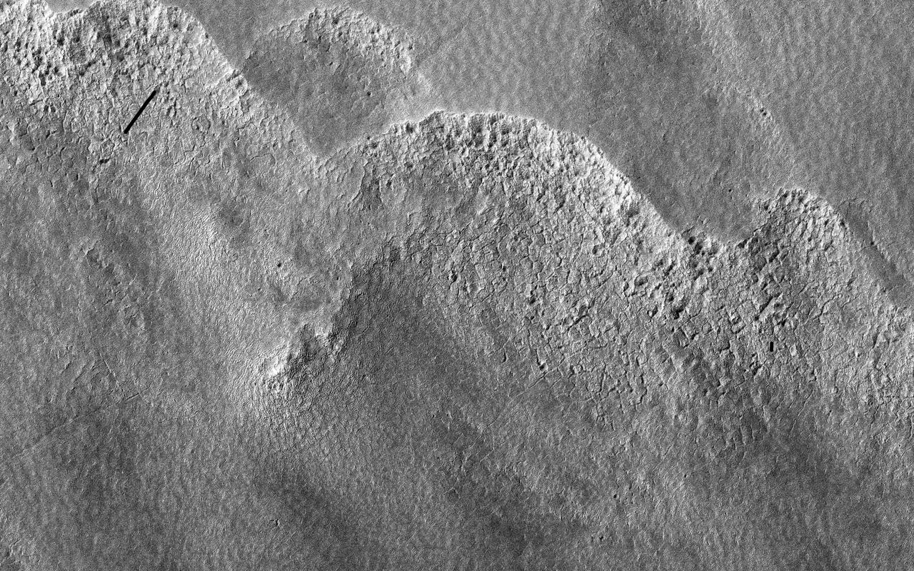 This image is of a portion of the Southern plains region within Hellas, the largest impact basin on Mars, with a diameter of about 2300 kilometers 1400 miles, as observed by NASA Mars Reconnaissance Orbiter. There are three main phenomena apparent in this image. First, the faint dark streaks that criss-cross the terrain are dust devil tracks that clear the bright dust along their way. Second, the subtle overall bumpy "basketball" texture of the surface is formed by repeated seasonal freezing and thawing of the ice-rich regolith and is common at higher latitudes. Third, the large, elliptical, scalloped depressions are common in permafrost terrains in both hemispheres, where thick, latitude-dependent sedimentary mantles comprise the surface units. These mantles are composed of ice-rich sediments that degrade as the ice sublimates away and is heated either by the Sun or by locally higher geothermal gradients. Sublimation, or the direct change in phase from ice to gas, occurs on Mars because of its low density atmosphere. These depressions have steeper pole-facing slopes, whereas the equator-facing slopes gently fade into the surrounding terrain. At full resolution (see close up view), numerous sublimation pits and networks of polygonal cracks are visible on the steeper, unstable pole-ward facing slopes. The overall morphology of this terrain is characteristic of what is called "thermokarstic degradation processes," which is a term used to describe the formation of pits in an ice-rich terrain due to loss of ice creating pits and collapse features. http://photojournal.jpl.nasa.gov/catalog/PIA19350