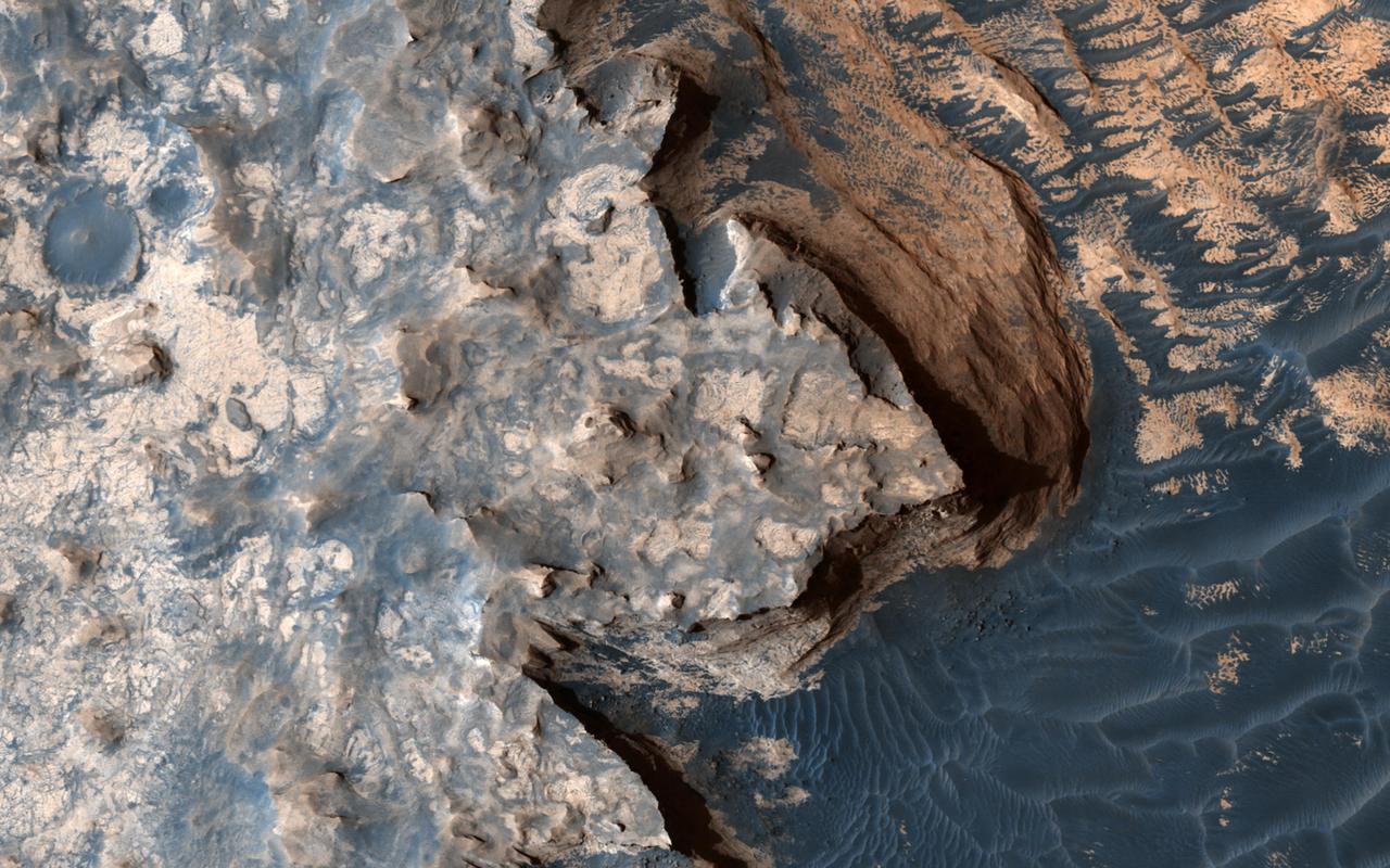 This image from NASA's Mars Reconnaissance Orbiter is reminiscent of the rugged and open terrain of a stark shore-line, perhaps of an island nation, such as the British Isles. A close-up in enhanced color produces a striking effect, giving the impression of a cloud-covered cliff edge with foamy waves crashing against it.  The reality is that the surface of Mars is much dryer than our imaginations might want to suggest. This is only a tiny part of a much larger structure; an inverted crater -- a crater that has been infilled by material that is more resistant to erosion than the rocks around it -- surrounded by bluish basaltic dunes. The edge of these elevated light-toned deposits are degraded, irregular and cliff-forming.  Dunes visible below the cliff, give the impression of an ocean surface, complete with foam capped waves crashing against the "shore line," demonstrating the abstract similarity between the nature of a turbulent ocean and a Martian dune field.  Meridiani Planum has an overall smooth terrain, which starkly contrasts with the more common boulder- and crater-laden landscapes observed over much of the rest of Mars. This makes it relatively younger in character than many other areas of the planet. Meridiani is one of the Mars Exploration Rover landing sites, and, is known for its layers and sediments. The orbital detection of hematite was one of the main reasons for sending Opportunity to this area.  Salt-bearing rocks -- also called sulphates -- were observed in the very first image from Opportunity, so perhaps it's apt that this HiRISE image reminds us of the turmoil and rugged beauty of a cliff-face, a coastline, being worn down by a relentless sea.   https://photojournal.jpl.nasa.gov/catalog/PIA21760