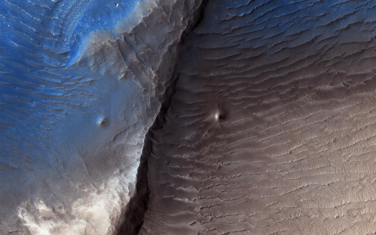 Geologists aren't quite sure what to make of the dark splotch in the middle of this image from NASA's Mars Reconnaisance Orbiter (MRO) -- one of several similar dark splotches that extend east and west for over 100 kilometers. From measurements made in infrared, this and other dark splotches have what we call "high thermal inertia," meaning that it heats up and cools down slowly. Scientists use thermal inertia to assess how rocky, sandy, or dusty a place is. A higher thermal inertia than the surrounding area means it's less dusty. Wavy, banded patterns in the dark splotch (possibly due to cross bedding from sand dunes that once occupied the area) were lithified into sandstone, and then eroded away. These clues could help geologists figure out what's going on there. https://photojournal.jpl.nasa.gov/catalog/PIA22042