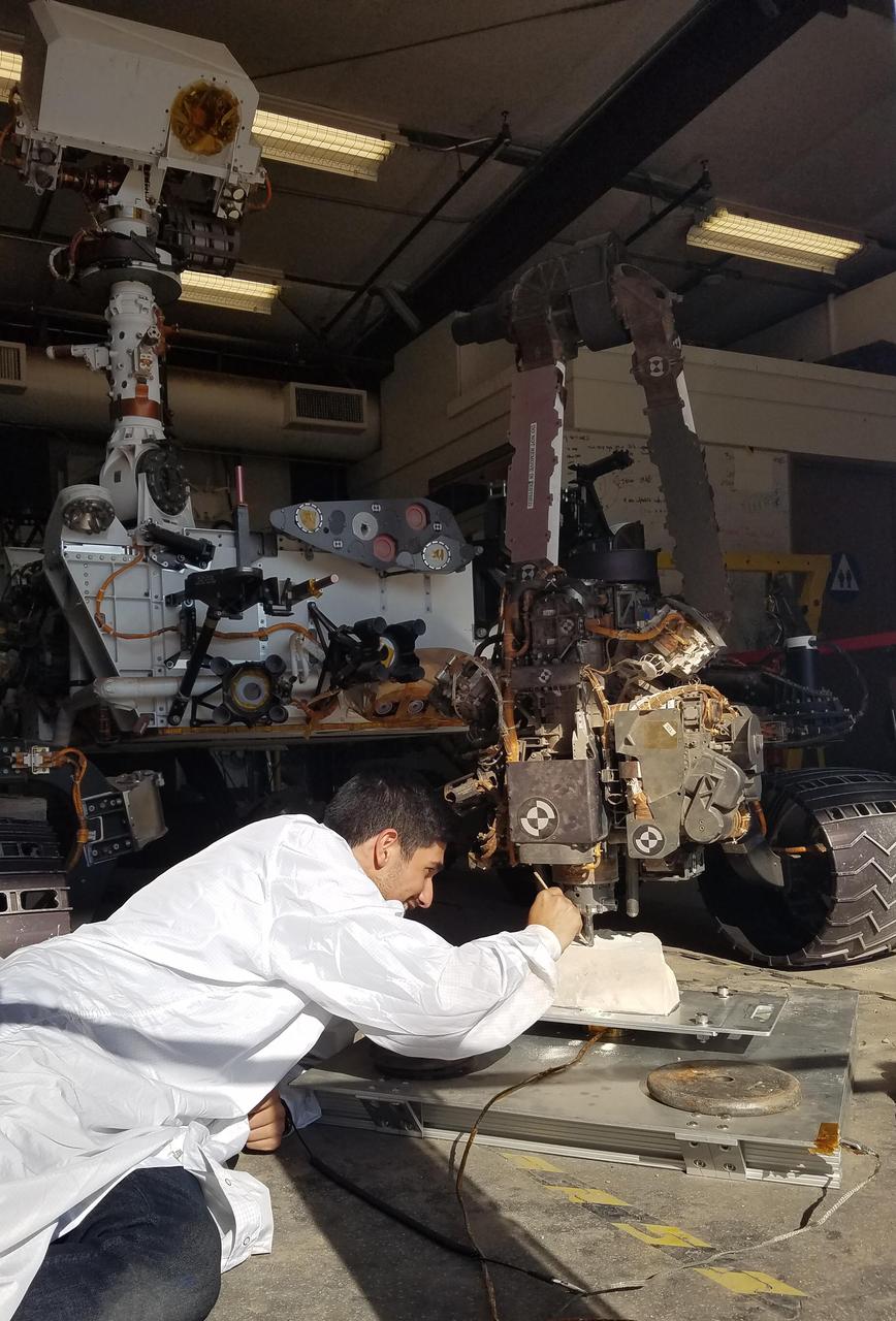 In the summer and fall of 2017, the team operating NASA's Curiosity Mars rover conducted tests in the "Mars Yard" at NASA's Jet Propulsion Laboratory, Pasadena, California, to develop techniques that Curiosity might be able to use to resume drilling into rocks on Mars.  JPL robotics engineer Vladimir Arutyunov, in this June 29, 2017, photo, checks the test rover's drill bit at its contact point with a rock. Note that the stabilizer post visible to the right of the bit is not in contact with the rock, unlike the positioning used and photographed by Curiosity when drilling into rocks on Mars in 2013 to 2016.  In late 2016, after Curiosity's drill had collected sample material from 15 Martian rocks, the drill's feed mechanism ceased working reliably. That motorized mechanism moved the bit forward or back with relation to the stabilizer posts on either side of the bit. In normal drilling by Curiosity, the stabilizers were positioned on the target rock first, and then the feed mechanism extended the rotation-percussion bit into the rock.  In the alternative technique seen here, called "feed-extended drilling," the test rover's stabilizers are not used to touch the rock. The bit is advanced into the rock by motion of the robotic arm rather than the drill's feed mechanism.   https://photojournal.jpl.nasa.gov/catalog/PIA22061