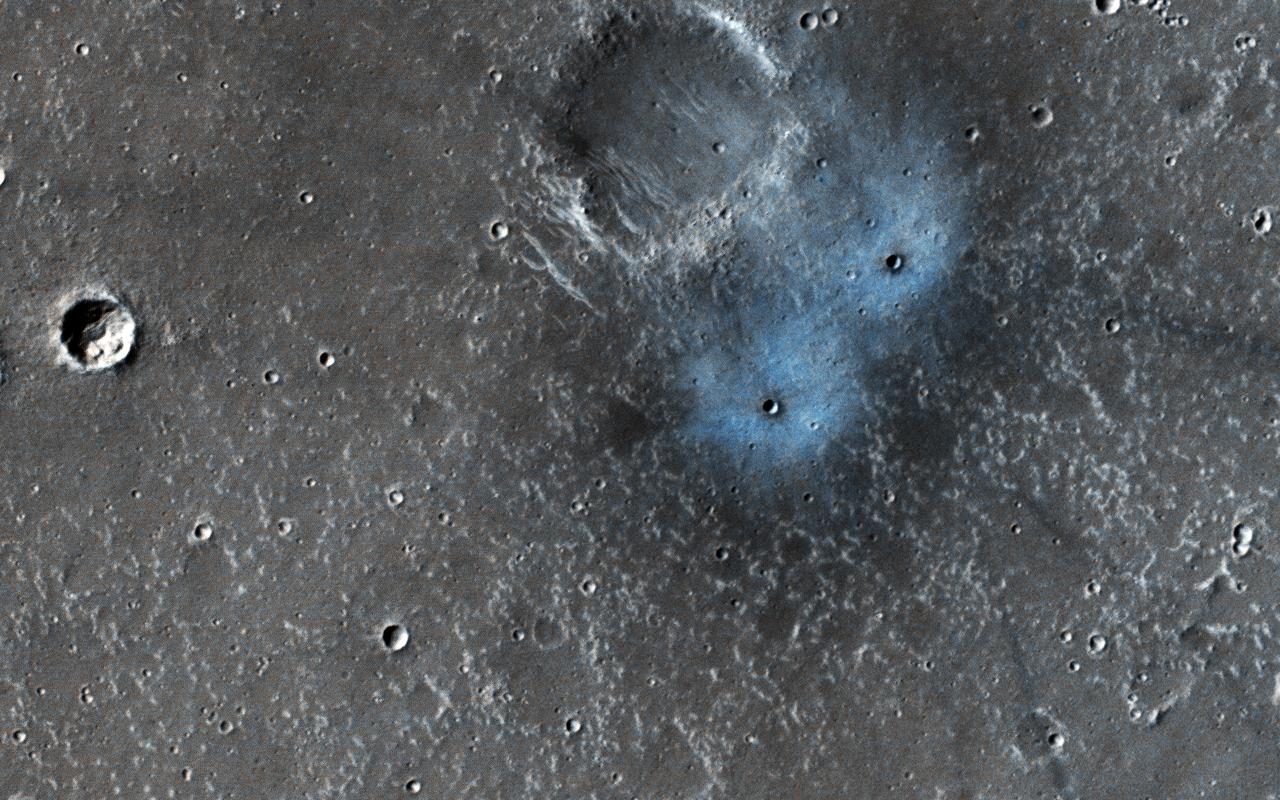 This new impact was found by NASA's Mars Reconnaissance Orbiter's Context Camera team (CTX), who asked HiRISE to take a high-resolution image. They estimated the impact happened between January 2012 and September 2016, because they have a picture from the THEMIS instrument without it before that time frame. These craters may be somewhere between two and four (Earth) years old, which is exceedingly young in geologic terms. Most of the craters we see on Mars (like others in this picture) are millions of years old. The blue appearance is due to the intense blast of the impact moving around dust on the surface. That dust is usually light-toned and reddish in color compared to what's beneath it. When you remove the dust, you're left with a dark spot that can sometimes be blue in comparison to the redder surroundings. These features are prominent only around young impacts, because all the old craters get covered with dust again over time. This blueish "blast zone" is very different than the exposed ice some new craters dig up. The shape, color, and brightness are distinct and tell scientists this is not ice. More information is available at https://photojournal.jpl.nasa.gov/catalog/PIA22534