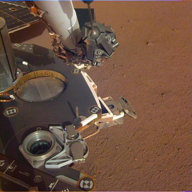 A partial view of the deck of NASA's InSight lander, where it stands on the Martian plains Elysium Planitia. The color-calibrated image was received on Dec. 4, 2018 (Sol 8). InSight's robotic arm with its stowed grapple can be seen above the deck, and jutting out from the front of the deck is one of the boxy attitude control system thrusters that helped control the spacecraft's landing. The circular silver inset of the propellant tank can also be seen in the middle of the image, as well as one of the connections for the aeroshell and parachute, which looks like a cupholder in the foreground. Next to the propellant tank is the UHF antenna, which helps the lander communicate with Earth. In the background, part of one of InSight's solar panels is visible. https://photojournal.jpl.nasa.gov/catalog/PIA22873