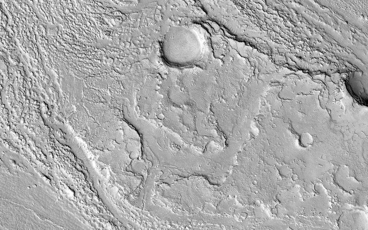 This image in Athabasca Valles shows lava flows originating from Elysium Mons to the northwest. A Context Camera image shows the lava flowed from the northwest to the southeast, diverting around obstacles as it settled. (The flow is outlined in blue with the flow direction shown in yellow, and the approximate location of the HiRISE image is represented by a white rectangle.) The lava appears to have flowed smoothly around obstructions, almost like water, forming streamlined islands. In the southern part of this image, a branch of the flow diverts around a small crater, and eventually rejoins the main part of the flow. Irregular-shaped ring structures appear on the northern end and are related to the volcanic activity that formed the flows. We also see a dense cluster of secondary craters that formed when material ejected from Corinto Crater (to the northwest) impacted the surface at high speed. At full-resolution, this terrain has the distinctive appearance of a field of numerous, small and closely-spaced craters. https://photojournal.jpl.nasa.gov/catalog/PIA23062