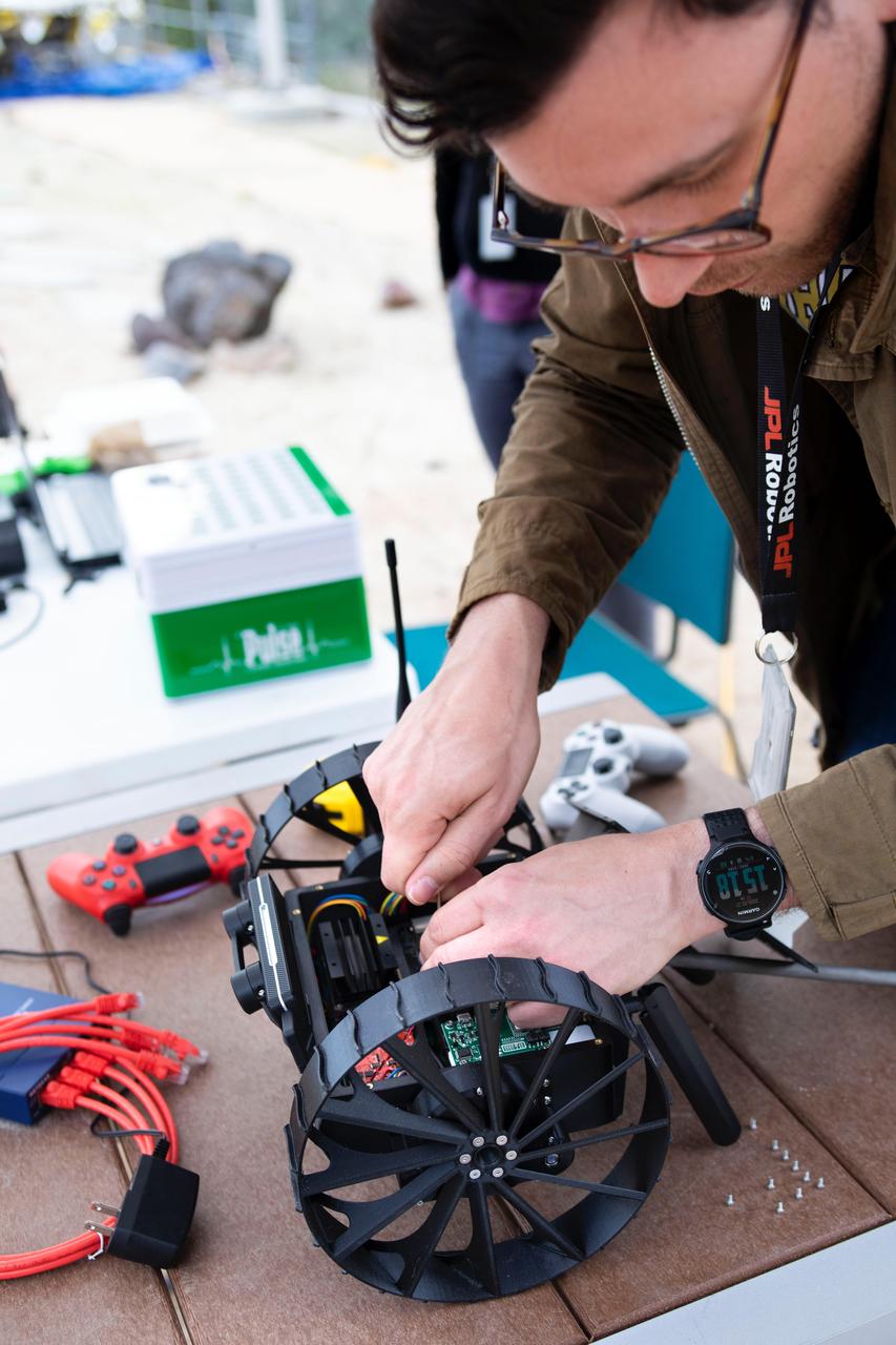Project manager Jean-Pierre de la Croix works on an Autonomous Pop-Up Flat Folding Explorer Robot (A-PUFFER) during recent trials in the Mars Yard at NASA's Jet Propulsion Laboratory. This PUFFER is equipped with an onboard computer and stereo camera as well as other sensors to help it autonomously navigate and collaborate with other PUFFERs.  https://photojournal.jpl.nasa.gov/catalog/PIA23794