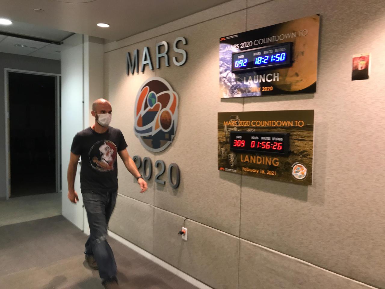 Ian Clark walks past mission countdown clocks in the Perseverance offices at NASA's Jet Propulsion Laboratory in Southern California. The Lab instituted a suite of safe@work procedures — based on the guidance of occupational safety medical personnel — to ensure those working at JPL are social distancing, wearing protective equipment and have ready access to hand sanitizer and other cleaning supplies during the coronavirus pandemic. Clark is one of a small subset of project personnel whose mission-essential job required physical access to the facility. He was on-Lab to supervise the assembly and cleaning of the sample tubes that will hold Martian sediment and rock for return to Earth on a future mission.  https://photojournal.jpl.nasa.gov/catalog/PIA23830