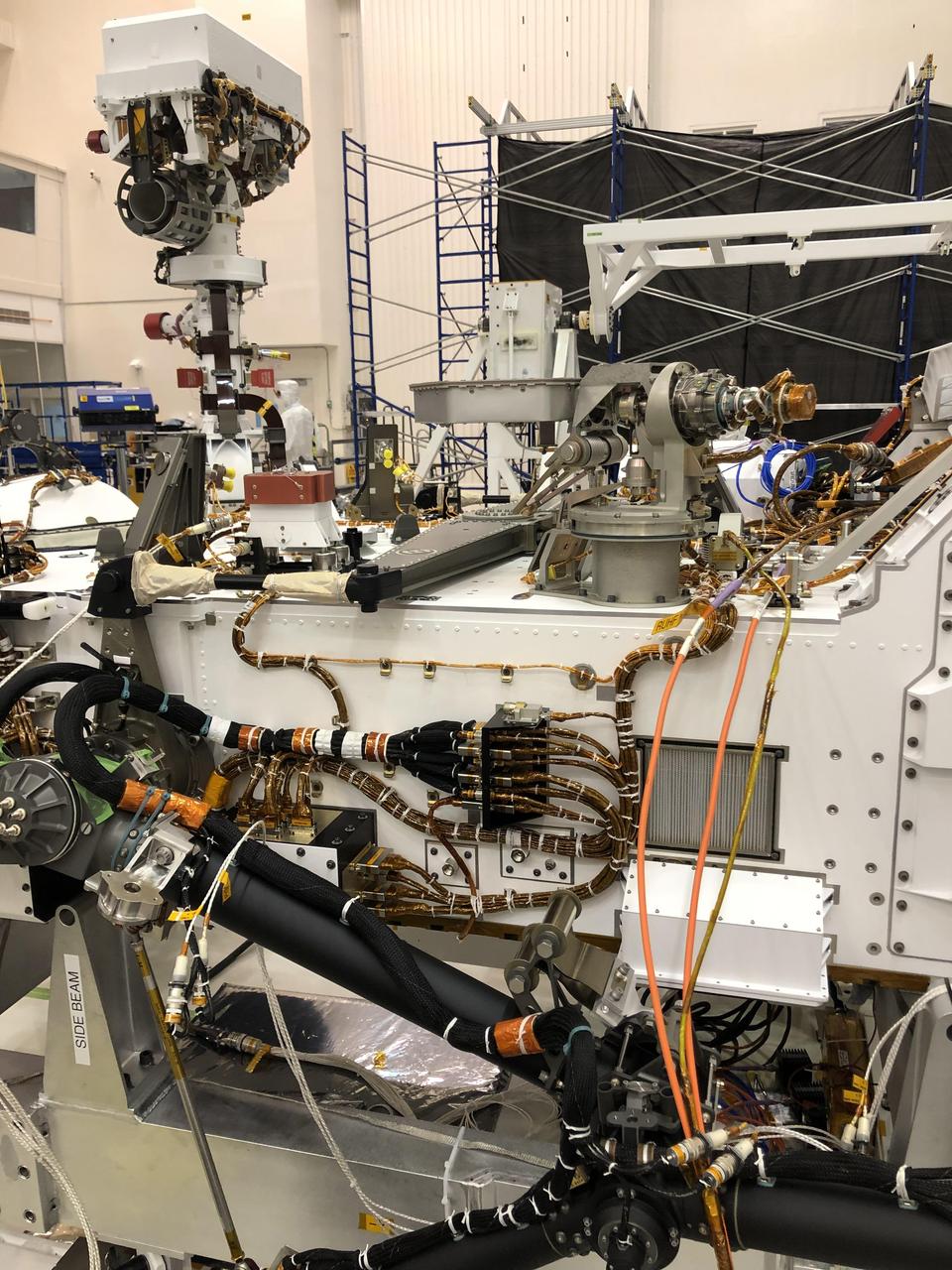 The port side of NASA's Perseverance Mars rover can be seen in this image taken on Nov. 16, 2019, in High Bay 1 of the Spacecraft Assembly Facility at NASA's Jet Propulsion Laboratory in Southern California. At the top left, the rover's remote sensing mast can be seen in the deployed position. To the right of the mast in the center of the image is the light gray high-gain antenna. At center of the image, attached to the side of the rover, is a black cable bracket (with gold cabling running through it). Attached to the top of this black bracket — and gray in color — is the Entry Descent and Landing (EDL) microphone. The rectangular screen to the right of the cable bracket is the rover chassis HEPA filter, which is above the white box housing the Mars Oxygen In-Situ Resource Utilization Experiment (MOXIE) inlet filter assembly. Gray and bright orange cables seen in the foreground of the image belong to ground support equipment.  https://photojournal.jpl.nasa.gov/catalog/PIA24045