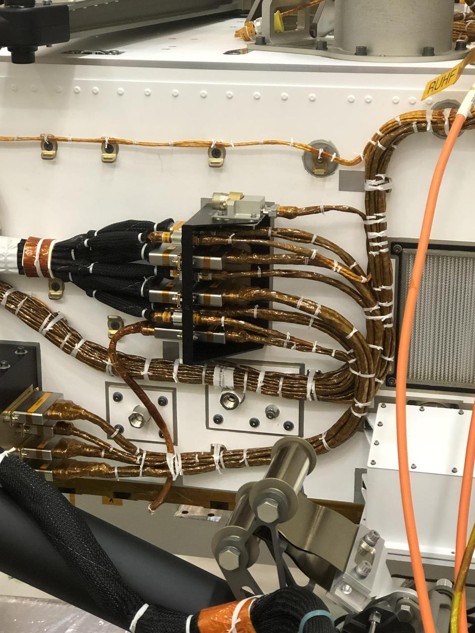 Components are visible on the port side of the Perseverance rover in this close-up image taken on Nov. 16, 2019, in High Bay 1 of the Spacecraft Assembly Facility at NASA's Jet Propulsion Laboratory in Southern California. At center of the image, attached to the side of the rover, is a black cable bracket (with gold cabling running through it). Attached to the top of this black bracket — and gray in color — is the Entry Descent and Landing (EDL) microphone. Below the cable bracket are the Mars Oxygen In-Situ Resource Utilization Experiment (MOXIE) wall ports. The orange cable passing over it is part of ground support equipment.  https://photojournal.jpl.nasa.gov/catalog/PIA24046