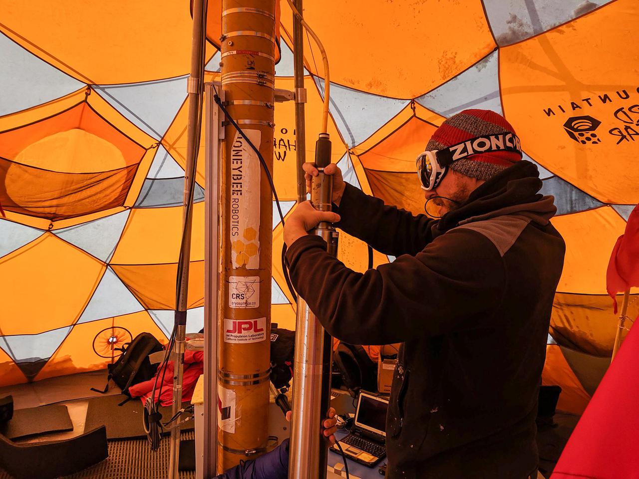 During 2019 field tests near Greenland's Summit Station, a high-elevation remote observing station, the WATSON (Wireline Analysis Tool for the Subsurface Observation of Northern ice sheets) instrument is put through its paces to seek out signs of life, or biosignatures, 360 feet (110 meters) down a borehole. In this photograph, a WATSON team member secures the tether to the top of the tube-like instrument and drill before lowering it into the ice. The tether also acts as the power cable and data feed. Care must be taken to ensure a tight connection between the tether and instrument, else the instrument could be lost in the ice.      WATSON could one day be launched aboard a robotic mission to seek out biosignatures on the ocean moons of Enceladus, Europa, or even Titan. The WATSON team hopes to test the instrument in a variety of cold locations on Earth to see how the distribution and variety of biosignatures change depending on where they are. By testing WATSON in different Earth analogs — areas on Earth that can stand in for those on other worlds — scientists would be able to better understand the chemical fingerprints of any biosignatures detected on other worlds.  https://photojournal.jpl.nasa.gov/catalog/PIA24170