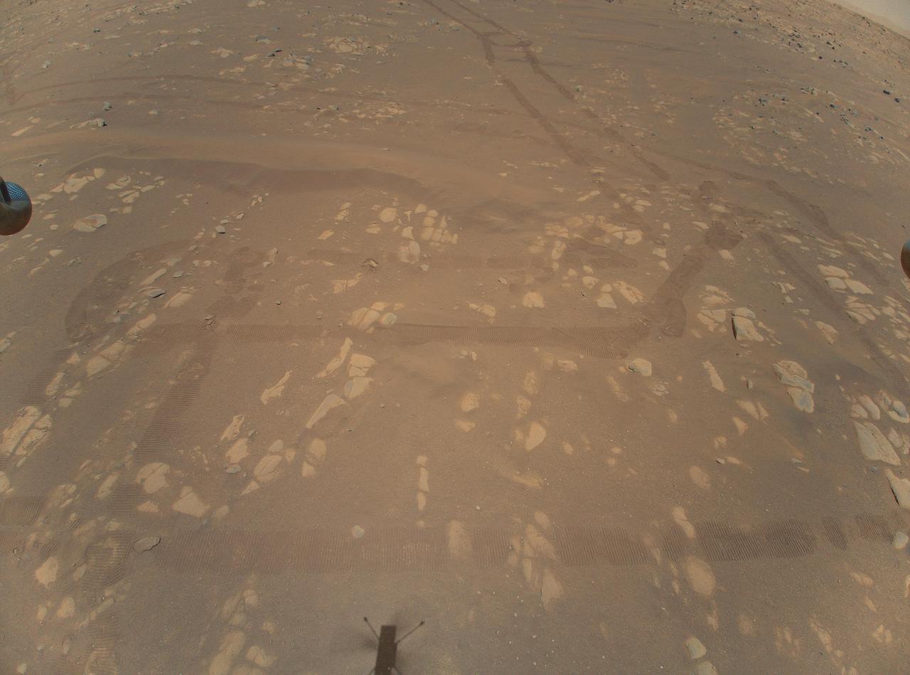 This is the first color image of the Martian surface taken by an aerial vehicle while it was aloft. The Ingenuity Mars Helicopter captured it with its color camera during its second successful flight test on April 22, 2021. At the time this image, Ingenuity was 17 feet (5.2 meters) above the surface and pitching (moving the camera's field of view upward) so the helicopter could begin its 7-foot (2-meter) translation to the west — away from the rover. The image demonstrates the utility of scouting Martian terrain from an aerial perspective.  The winding parallel discolorations in the surface reveal the tread of the six-wheeled rover. Perseverance itself is located top center, just out frame. "Wright Brothers Field" is in the vicinity of the helicopter's shadow, bottom center, with the actual point of takeoff of the helicopter just below the image. A portion of the landing pads on two of the helicopter's four landing legs can be seen in on the left and right sides of the image, and a small portion of the horizon can be seen at the upper right and left corners.  Mounted in the helicopter's fuselage and pointed approximately 22 degree below the horizon, Ingenuity's high-resolution color camera contains a 4208-by-3120-pixel sensor.   https://photojournal.jpl.nasa.gov/catalog/PIA24593