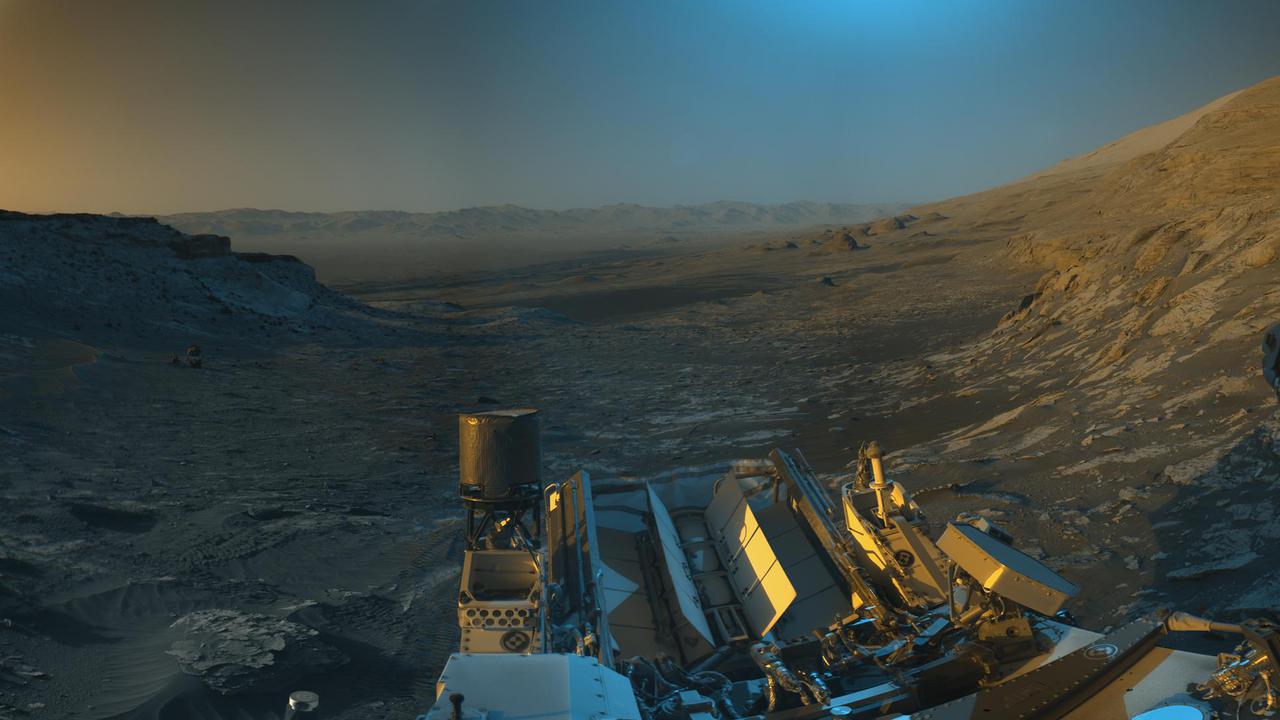 NASA's Curiosity Mars rover used its black-and-white navigation cameras to capture panoramas of this scene at two times of day. Blue, orange, and green color was added to a combination of both panoramas for an artistic interpretation of the scene.  On Nov. 16, 2021 (the 3,299th Martian day, or sol, of the mission), engineers commanded Curiosity to take two sets of mosaics, or composite images, capturing the scene at 8:30 a.m. and again at 4:10 p.m. local Mars time. The two times of day provided contrasting lighting conditions that brought out a variety of unique landscape details. They combined the two scenes in an artistic re-creation that includes images from the morning scene in blue, the afternoon scene in orange, and a combination of both in green.  The main image is an artistic interpretation of the scene. Figure 1 is the mosaic taken in the afternoon. Figure 2 is the mosaic taken in the morning.  At the center of the image is the view back down Mount Sharp, the 3-mile-tall (5-kilometer-tall) mountain that Curiosity has been driving up since 2014. Rounded hills can be seen in the distance at center-right; Curiosity got a closer view of these back in July, when the rover started to see intriguing changes in the landscape. A field of sand ripples known as the "Sands of Forvie" stretches a quarter- to a half-mile (400 to 800 meters) away.  At the far right of the panorama is the craggy "Rafael Navarro Mountain," named after a Curiosity team scientist who passed away earlier this year. Poking up behind it is the upper part of Mount Sharp, far above the area Curiosity is exploring. Mount Sharp lies inside Gale Crater, a 96-mile-wide (154-kilometer-wide) basin formed by an ancient impact; Gale Crater's distant rim stands 7,500 feet tall (2.3 kilometers), and is visible on the horizon about 18 to 25 miles away (30 to 40 kilometers).  https://photojournal.jpl.nasa.gov/catalog/PIA24937
