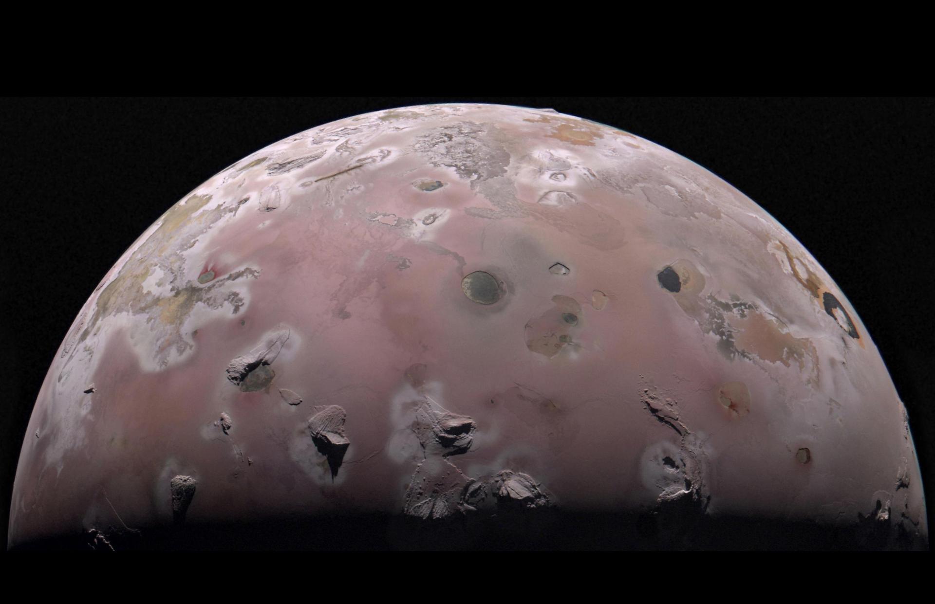 A view of half of the sphere of Jupiter’s volcanic moon Io against black space. The surface is colorful pinkish-tan and mottled with dark brownish features and sharp peaks surrounded by bright white diffuse deposits.