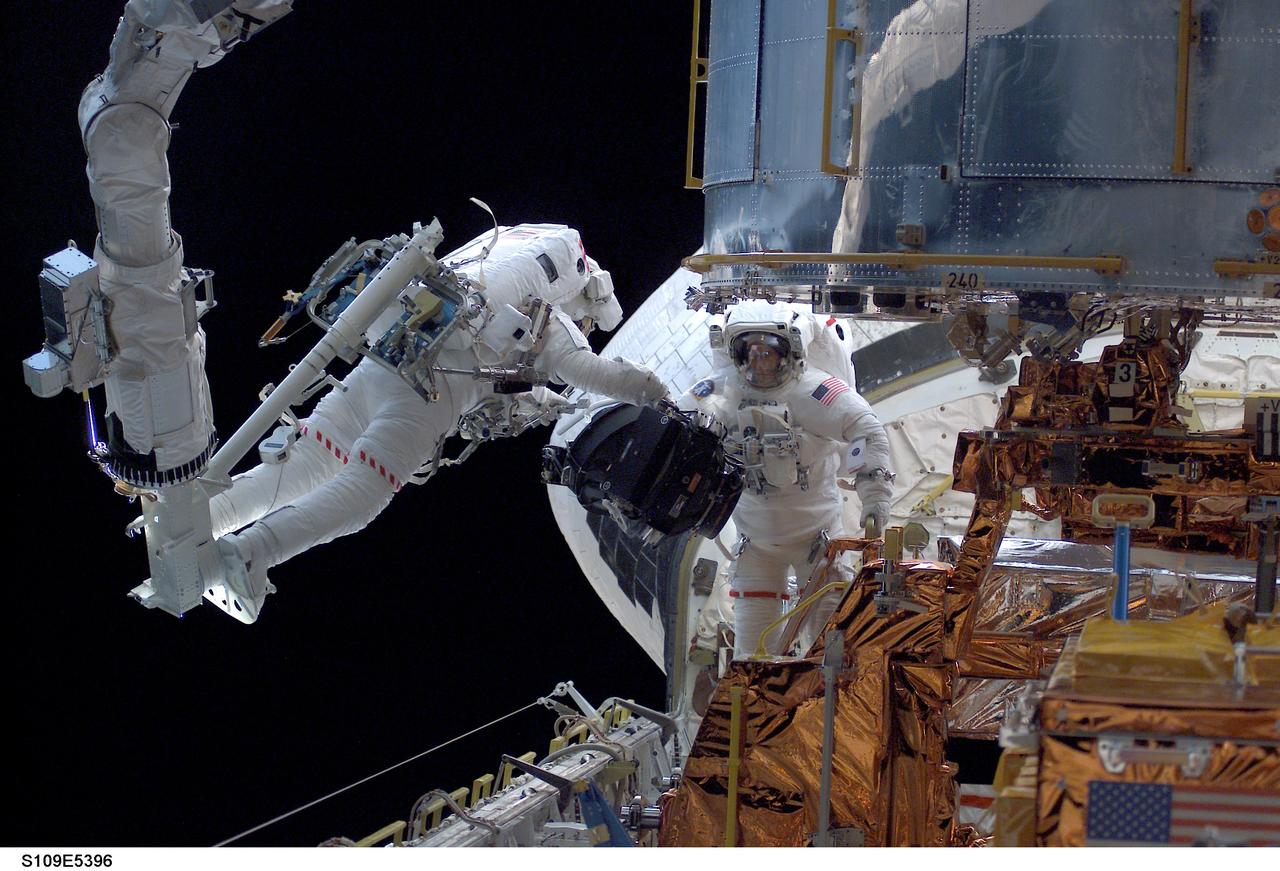 With his feet secured on a platform connected to the remote manipulator system (RMS) robotic arm of the Space Shuttle Columbia, astronaut Michael J. Massimino, mission specialist, hovers over the shuttle's cargo bay while working in tandem with astronaut James H. Newman, mission specialist, to replace the Reaction Wheel Assembly in the Hubble Space Telescope (HST) during the STS-109 mission's second day of extravehicular activity (EVA).