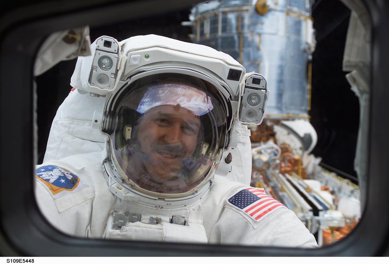 STS109-E-5448 (4 March 2002) --- Astronaut John M. Grunsfeld, payload commander, peers into the crew cabin of the Space Shuttle Columbia during the first STS-109 extravehicular activity (EVA-1) on March 4, 2002.  Grunsfeld's helmet visor displays a mirrored image of the Earth's hemisphere.  Astronauts Grunsfeld and Richard M. Linnehan replaced the starboard solar array on the Hubble Space Telescope (HST) on the first of five scheduled STS-109 space walks. The lower portion of the giant telescope can be seen over Grunsfeld's  left shoulder. The image was recorded with a digital still camera by a crewmate on shuttle's aft flight deck.