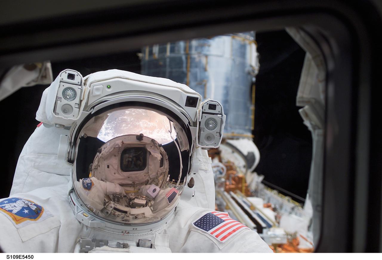 STS109-E-5450 (4 March 2002) --- Astronaut John M. Grunsfeld, payload commander, peers into the crew cabin of the Space Shuttle Columbia during the first STS-109 extravehicular activity (EVA-1) on March 4, 2002. Grunsfeld's helmet visor, with the sunshield now in place, displays mirrored images of the Earth's hemisphere and the Space Shuttle Columbia's aft cabin. The distorted reflection gives the crew cabin a cyclops-like appearance. Astronauts Grunsfeld and Richard M. Linnehan replaced the starboard solar array on the Hubble Space Telescope (HST) on the first of five scheduled STS-109 space walks. The lower portion of the giant telescope can be seen behind the payload commander. The image was recorded with a digital still camera by a crewmate on shuttle's aft flight deck.