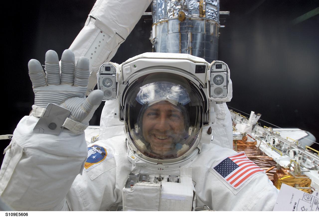 STS109-E-5606 (5 March 2002) --- Astronaut Michael J. Massimino, mission specialist, waves to crewmates on the other side of the aft flight deck windows on Columbia, while equipped with his extravehicular mobility units (EMU) space suit and standing on the end of the Remote Manipulator System (RMS) arm in the shuttle's cargo bay. This day's space walk went on to see astronauts James H. Newman and Massimino replace the port solar array on the Hubble Space Telescope (HST), partially visible in the background. On the previous day astronauts John M. Grunsfeld and Richard M. Linnehan replaced the starboard solar array on the giant telescope. The image was recorded with a digital still camera.