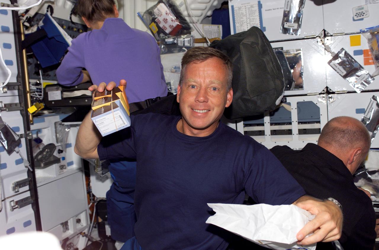 S121-E-05896 (7 July 2006) --- Astronaut Steven W. Lindsey, STS-121 commander, holds small boxes of food items on the middeck of the Space Shuttle Discovery. Also visible in the background are astronauts Lisa M. Nowak (left), mission specialist, and Mark E. Kelly, pilot.