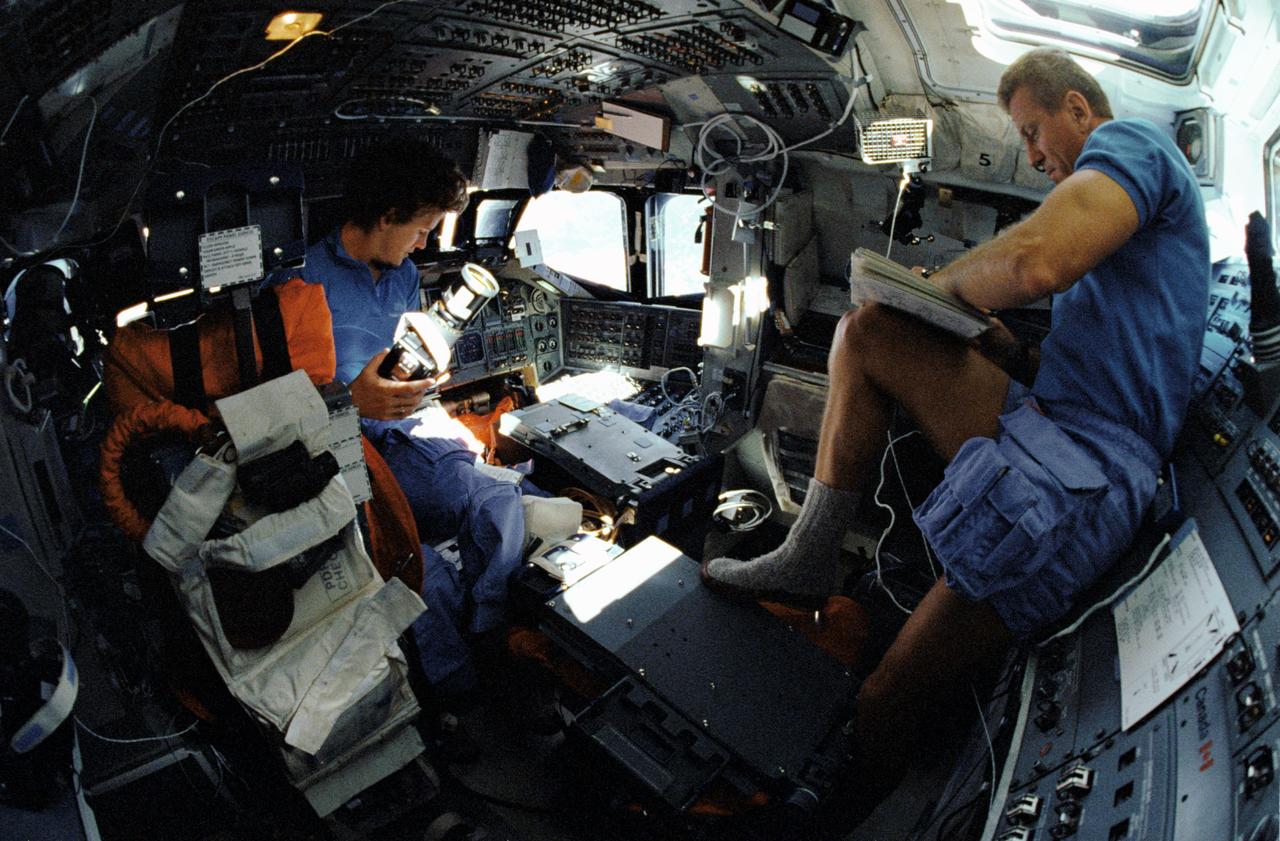 STS031-101-053 (24-29 April 199) --- A 35mm camera equipped with a "fish-eye" lens captured this view on Discovery's flight deck featuring astronaut Kathryn D. Sullivan with a Hasselblad camera on forward flight deck and astronaut Loren J. Shriver, pen in hand, amending flight data on aft flight deck.