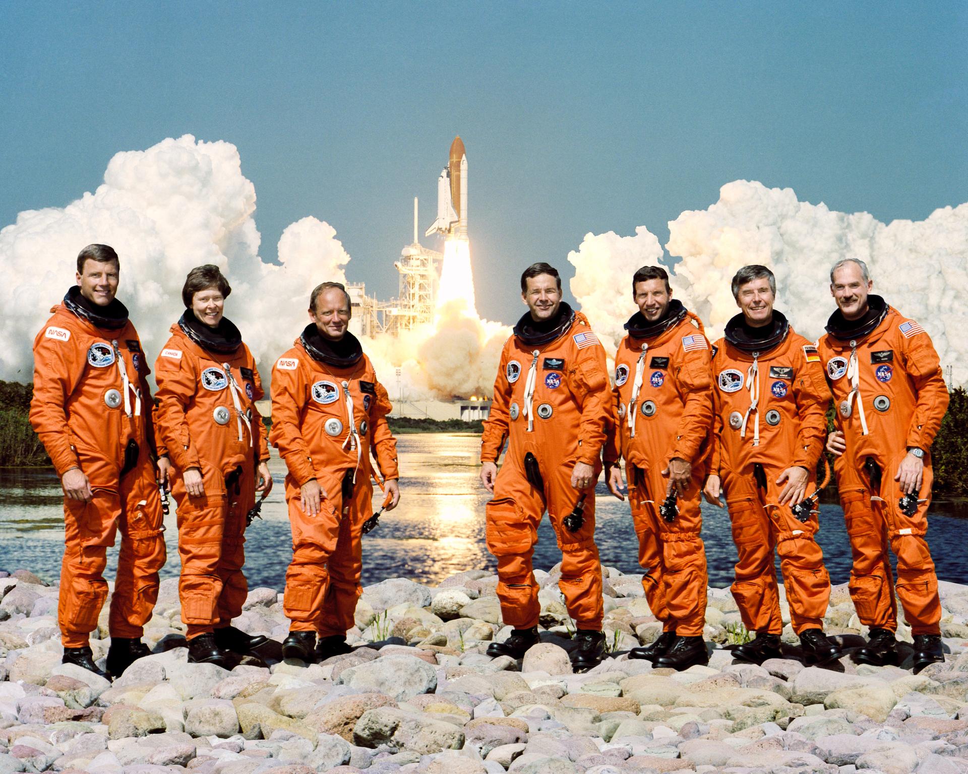 Payload specialists and astronauts (six men, one woman) pose for crew photo on rocks with shuttle taking off in background.