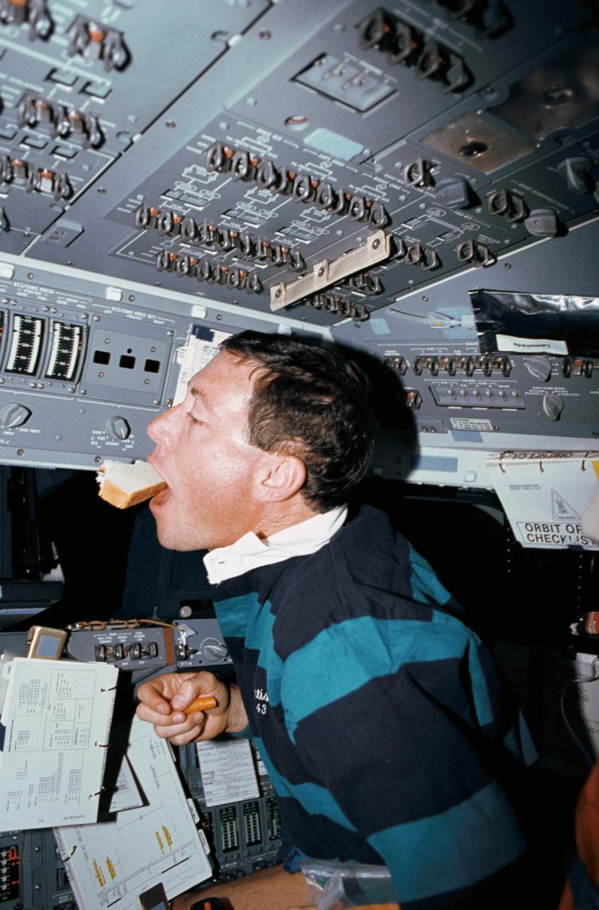 STS043-02-020 (2-11 Aug. 1991) --- Astronaut Michael A. Baker, STS-43 pilot, seated at the forward flight deck pilot station controls of the Space Shuttle Atlantis, eats a free-floating peanut butter and jelly sandwich while holding a carrot. Surrounding Baker are procedural checklists, control panels, and windows. A lemonade drink bag is velcroed to overhead panel.