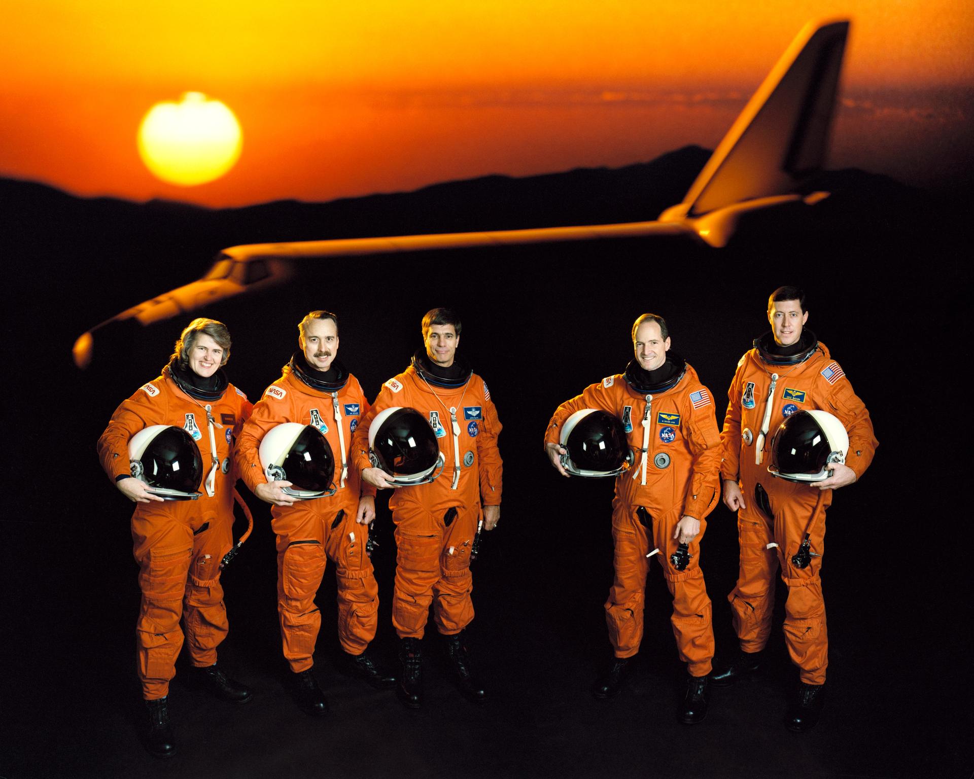 Five astronauts in orange space suit pose for crew photo in front of a shuttle and sunset backdrop.