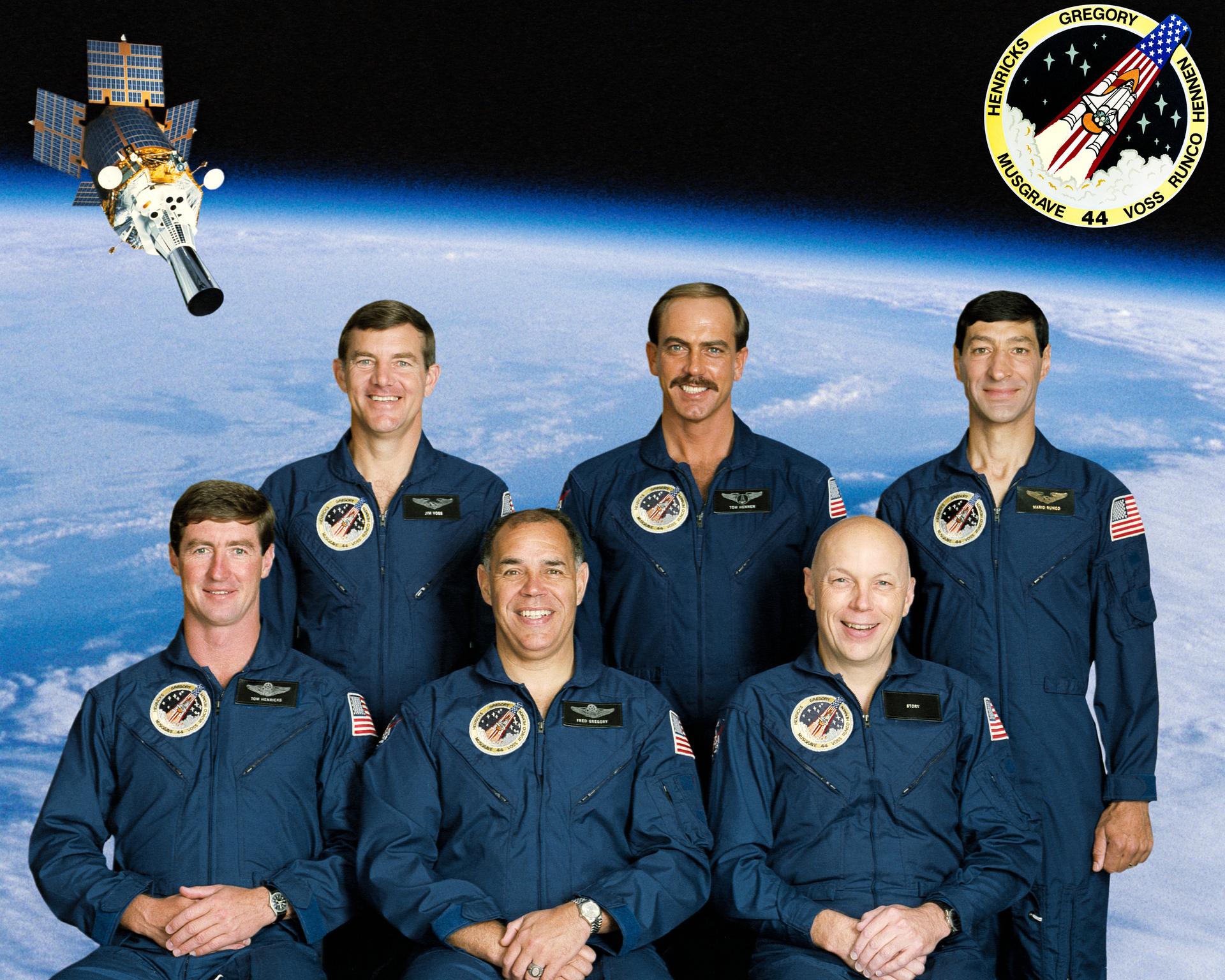 Six crew members in blue suits pose in front of an earth and satellite backdrop for crew photo.